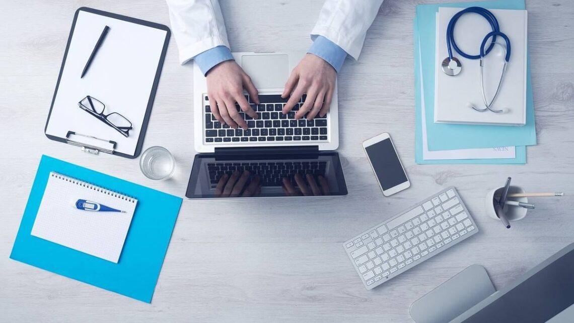 Main d'un médecin sur un ordinateur portable avec un stéthoscope et des documents médicaux à proximité
