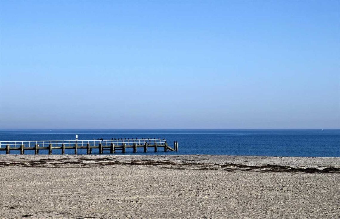 Plage sablonneuse, jetée en bois s'étendant sur la mer calme sous un ciel clair