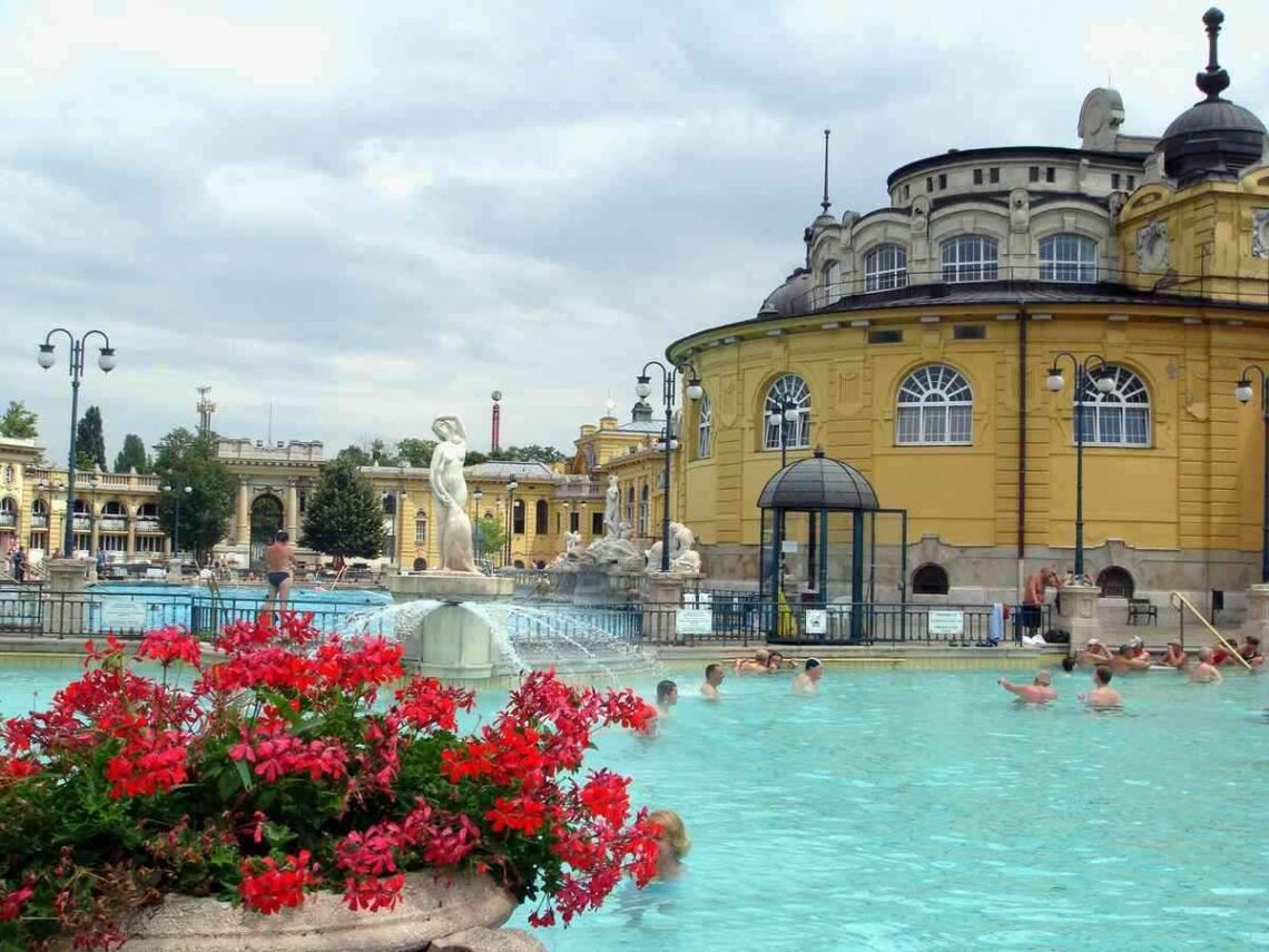 Piscine thermale turquoise entourée d'architecture jaune, baigneurs, fleurs rouges, bâtiments historiques
