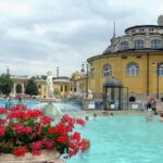 Piscine thermale turquoise entourée d'architecture jaune, baigneurs, fleurs rouges, bâtiments historiques