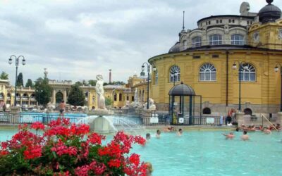 Piscine thermale turquoise entourée d'architecture jaune, baigneurs, fleurs rouges, bâtiments historiques