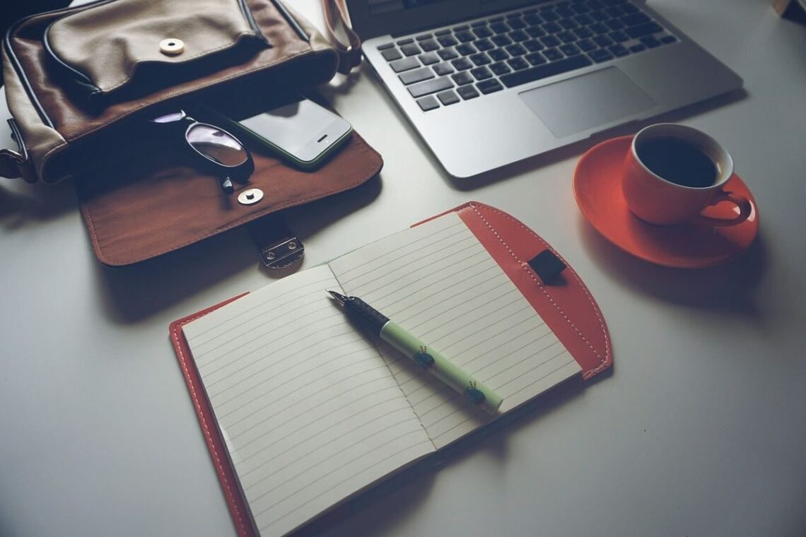 Un ordinateur portable, une tasse de café sur une soucoupe, un agenda ouvert avec un stylo, et un portefeuille sur une table