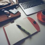 Un ordinateur portable, une tasse de café sur une soucoupe, un agenda ouvert avec un stylo, et un portefeuille sur une table